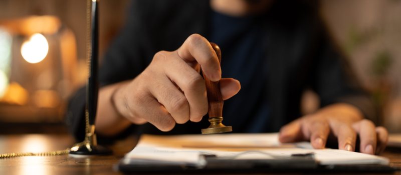 Close-up of a person's hand stamping with approved stamp on certificate document public paper at desk, notary or business people work from home, isolated for coronavirus COVID-19 protection